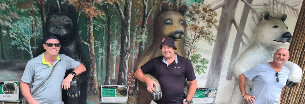 Three men posing beside bear sculptures in a forest setting, with informational plaques visible.