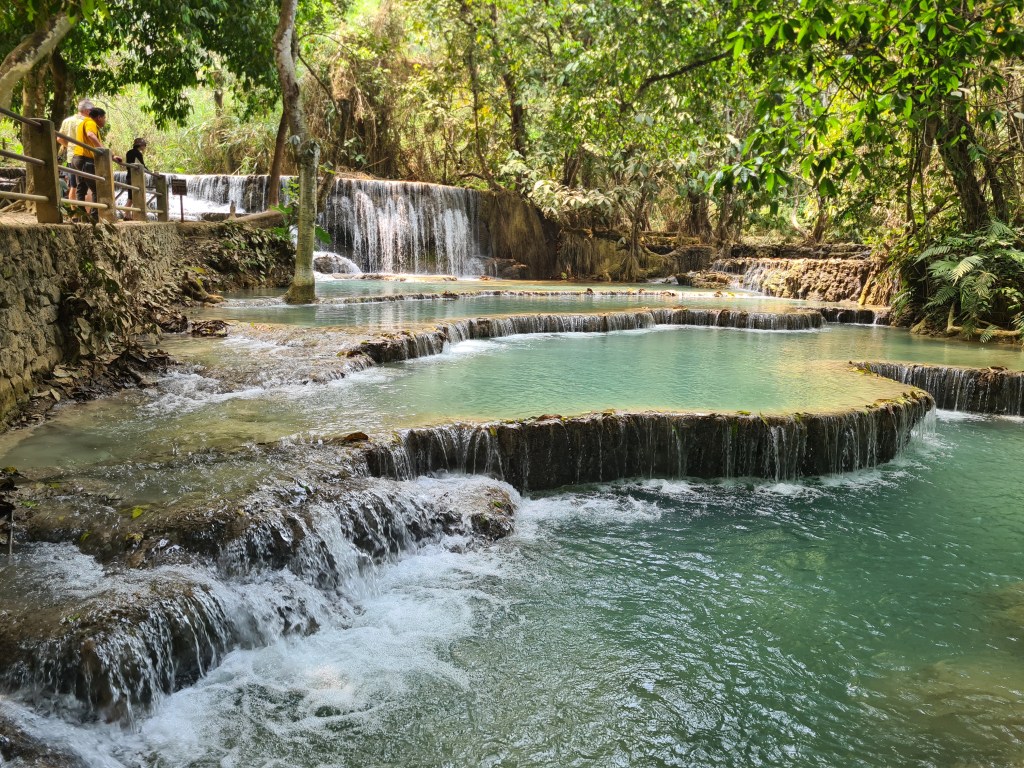 Lush green landscape featuring tiered waterfalls cascading into turquoise pools, with people enjoying the natural scenery.
