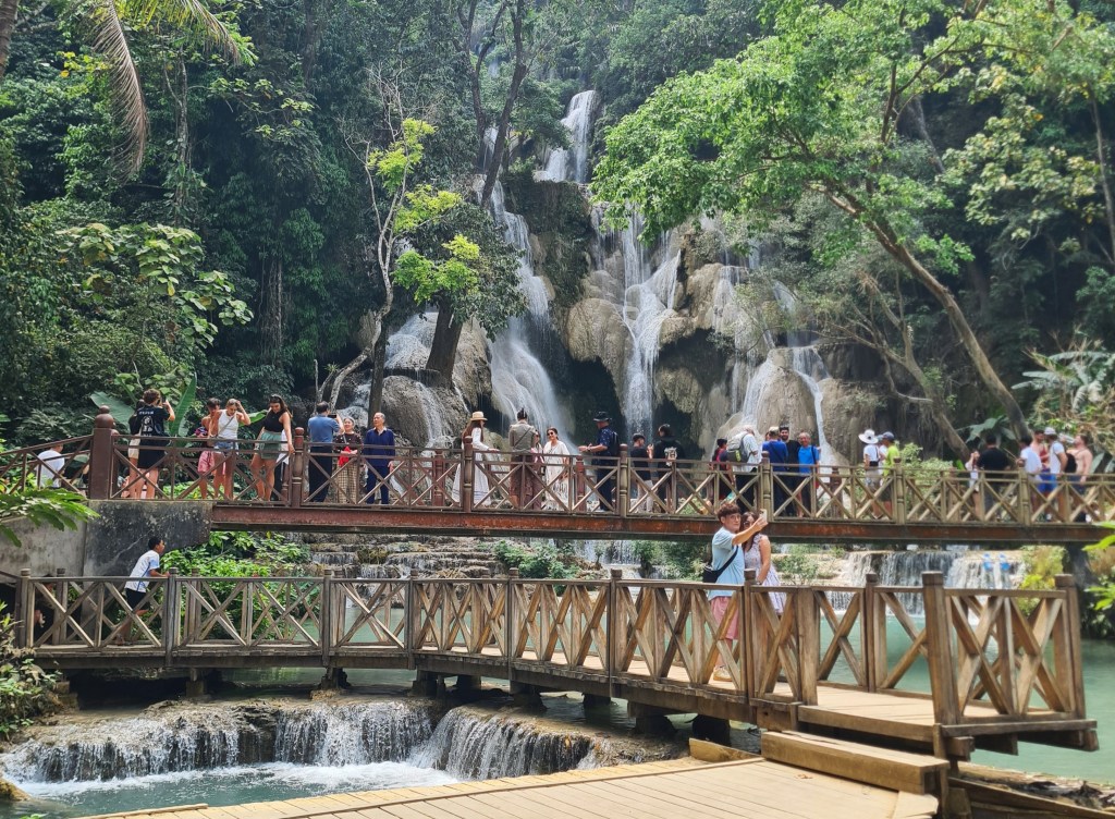People enjoying a visit to a scenic waterfall surrounded by lush greenery, with wooden bridges crossing over turquoise waters.