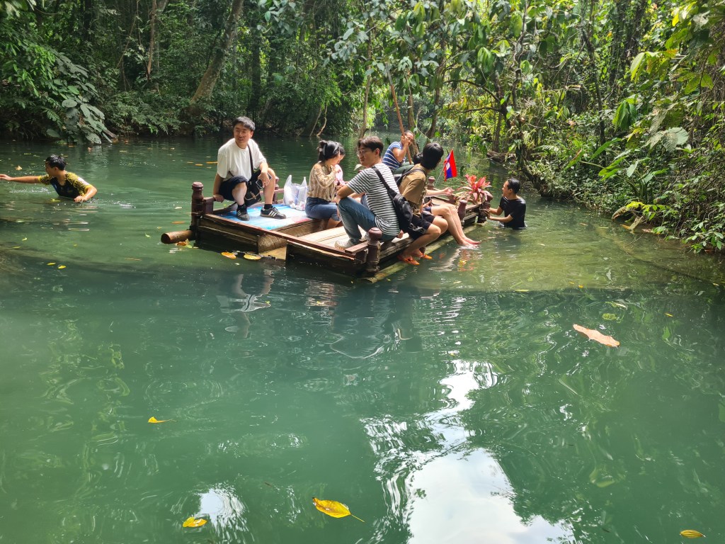 A group of people relaxing on a wooden float in a serene green river, with lush vegetation surrounding them. Some are sitting on the float, while others are swimming in the water.