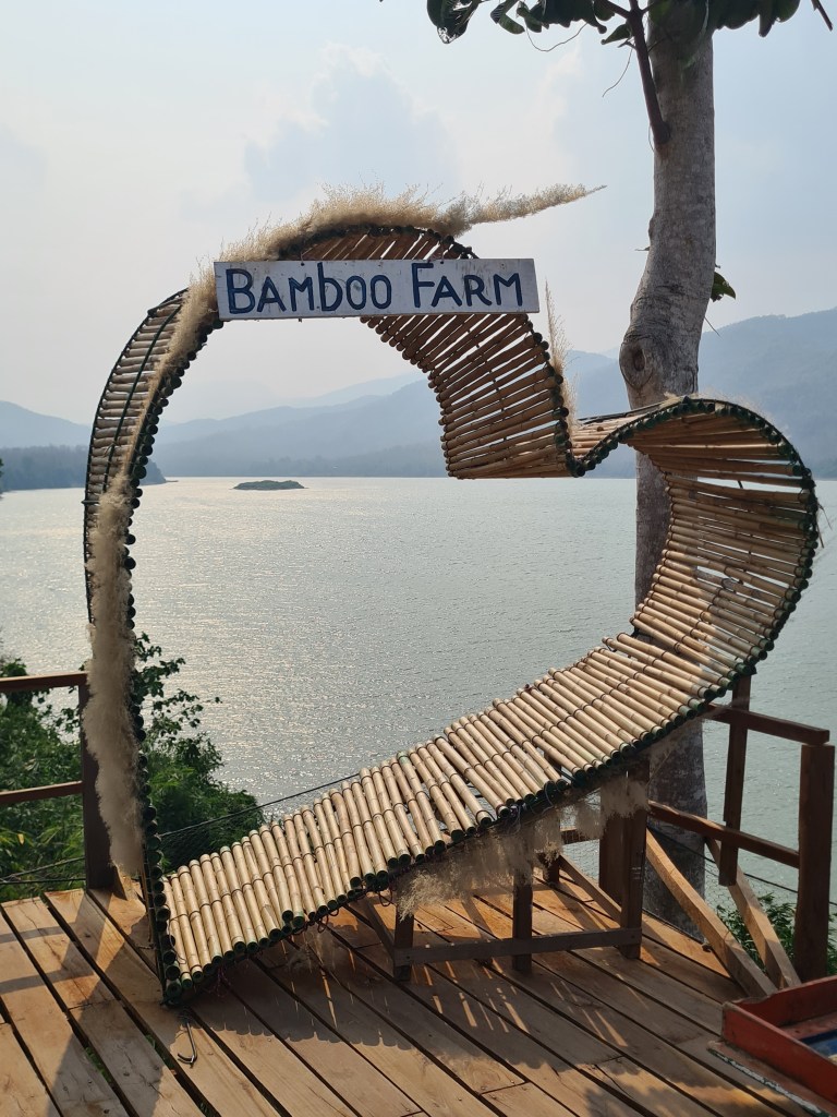 A decorative bamboo structure shaped like a heart, featuring the sign 'Bamboo Farm,' situated near a lake with mountains in the background.
