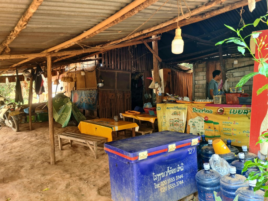 A rustic outdoor market stall with a wooden structure, featuring a display of goods including beverages and snacks. A person is standing behind the counter, while tables and chairs are set up for visitors. The area is surrounded by greenery and has a dirt floor.