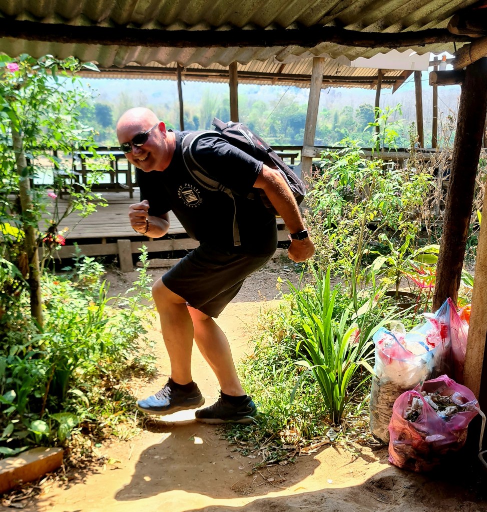 A man posing with a backpack, wearing sunglasses and a black t-shirt, is playfully stepping outside through a natural pathway surrounded by greenery and colorful flowers.