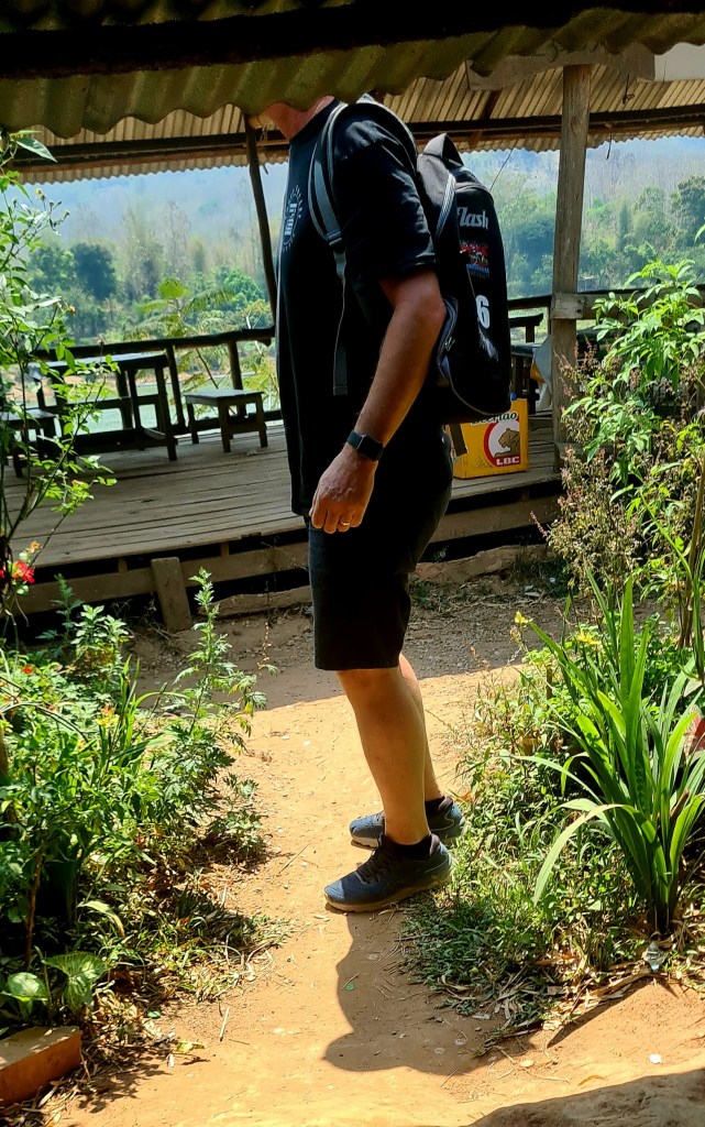 A person walking along a pathway surrounded by greenery and flowers, wearing a black t-shirt, shorts, and a backpack.