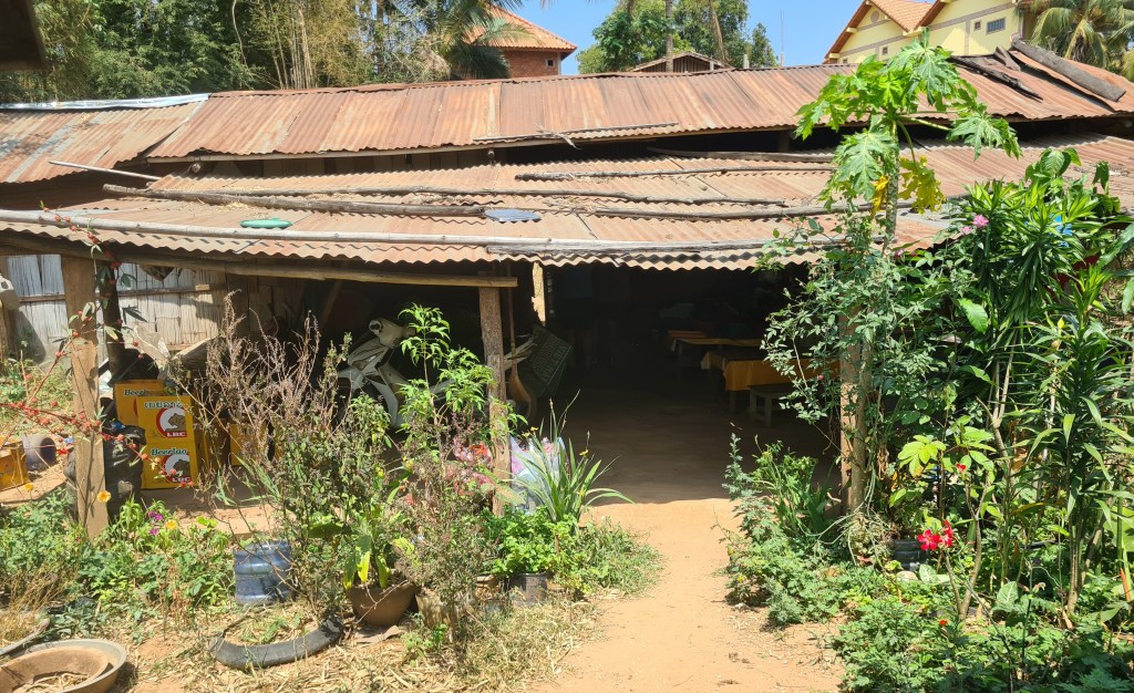 An old, rustic building with a corrugated metal roof surrounded by greenery and plants, featuring a dirt pathway leading to the entrance.