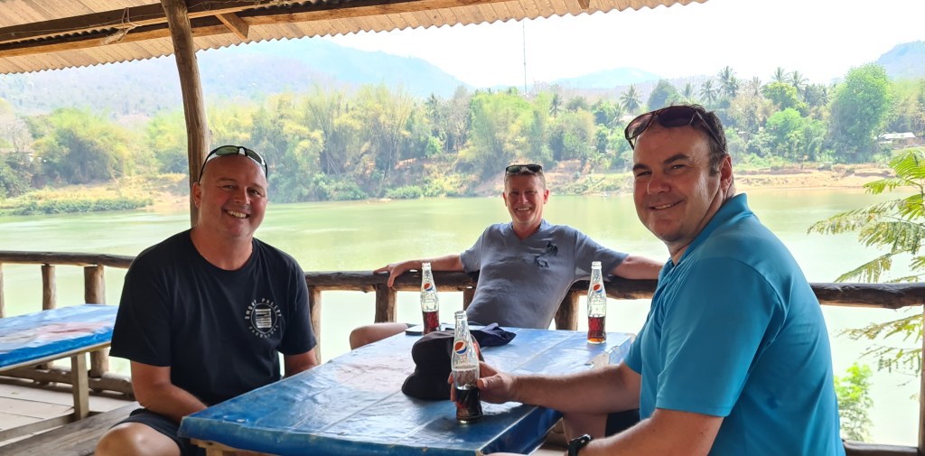 Three men sitting at a table with drinks, smiling and enjoying a scenic view of a river and lush greenery in the background.
