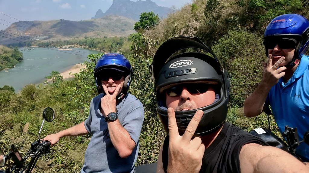 Three men wearing helmets making playful gestures while on motorcycles with a scenic river and mountains in the background.