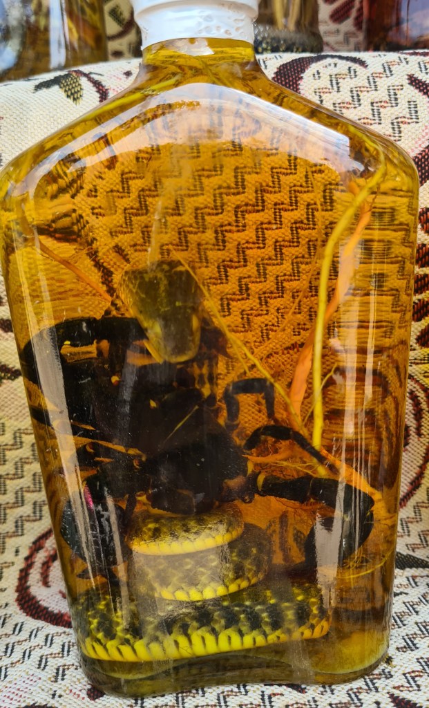 A glass bottle containing a mixture of preserved animals, including a snake and a scorpion, submerged in a golden liquid, placed on a patterned textile surface.