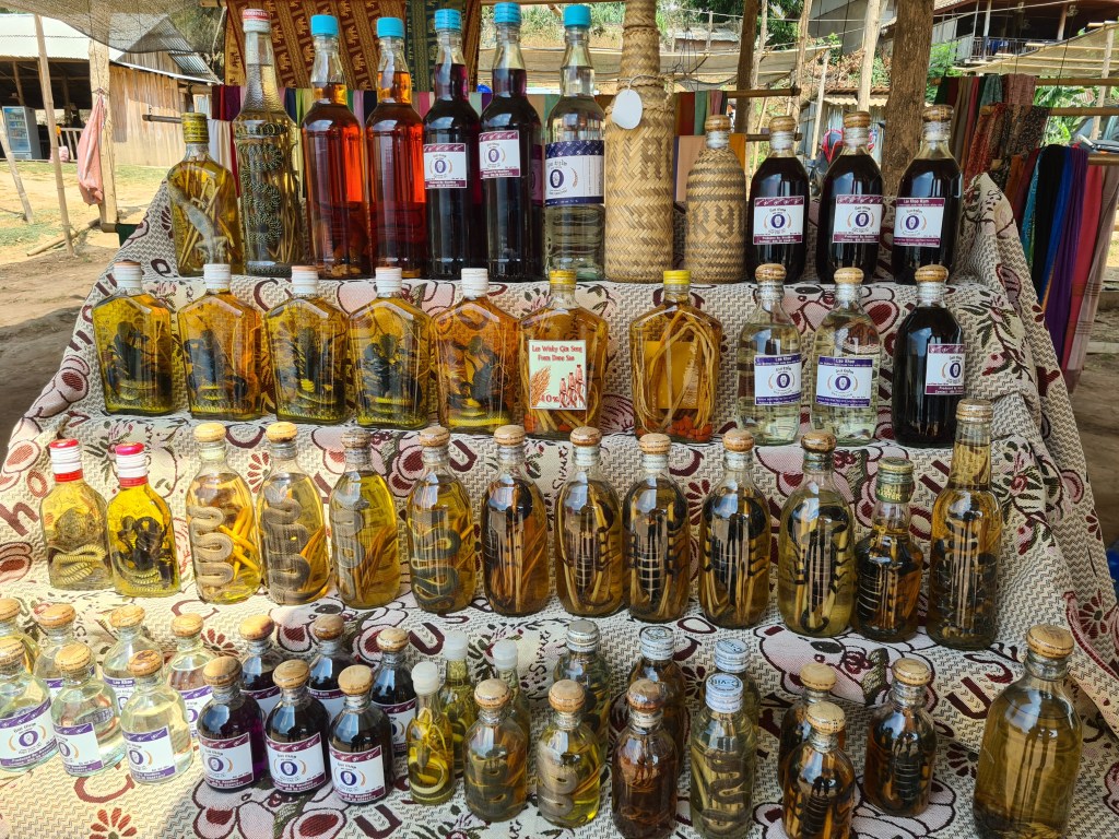 A variety of bottles containing different colored liquids, including several with snakes and other creatures, arranged on a patterned fabric. The display includes labels indicating the contents.