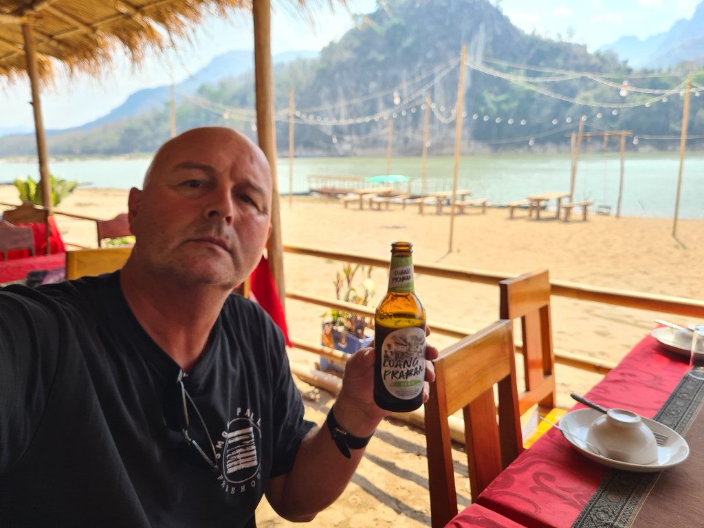 A man sitting at a table with a bottle of beer, overlooking a sandy beach and mountains in the background.