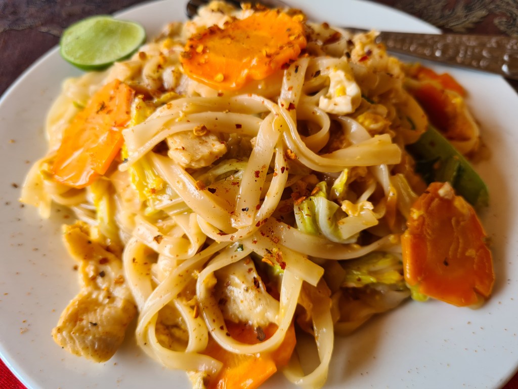 A close-up view of a plate of stir-fried noodles with vegetables, including carrots and green peppers, garnished with lime slices and sprinkled with red pepper flakes.