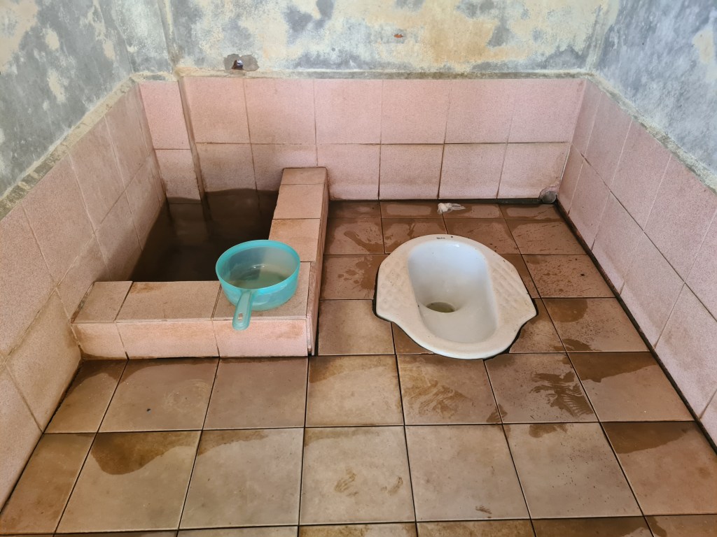 An indoor bathroom with a dirty tiled floor, featuring a squat toilet and a water basin sitting next to a shallow pool of water.