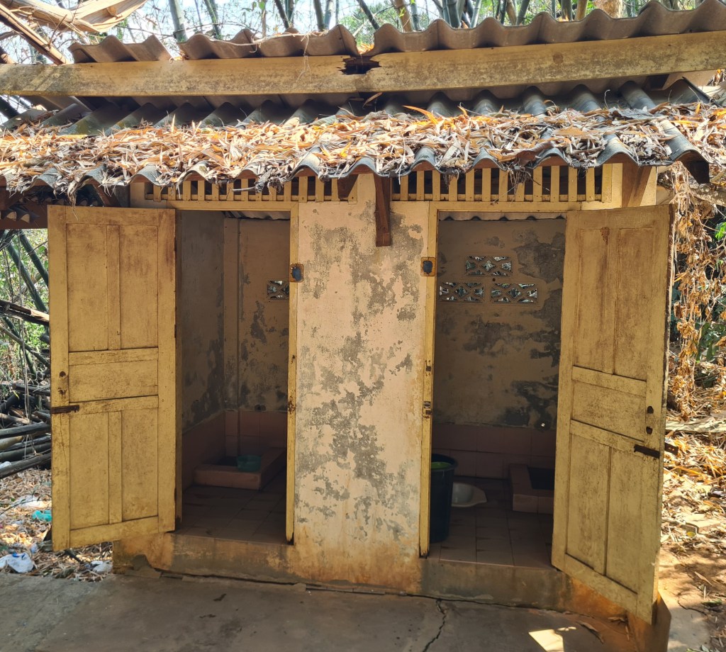 An old outdoor restroom building featuring three worn wooden doors, with a partially covered thatched roof and dry leaves on top.