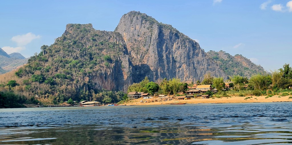 A scenic view of a riverbank with a small village nestled at the foot of a large, rocky mountain under a clear blue sky.