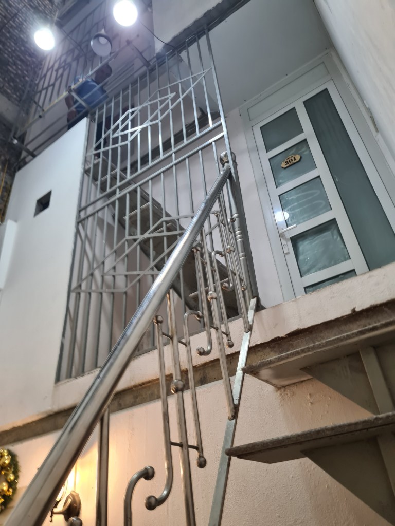 A staircase leading to a secured doorway, with a person working above on a platform, surrounded by metal railings and lighting.