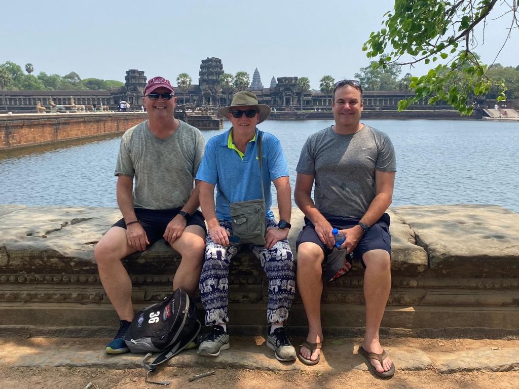 Three men sitting on a stone wall by a body of water, with ancient temple structures visible in the background.