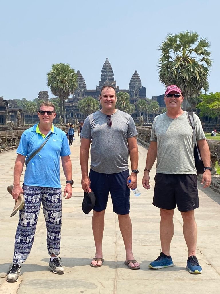 Three men standing on a pathway in front of Angkor Wat, with palm trees and temple structures visible in the background.