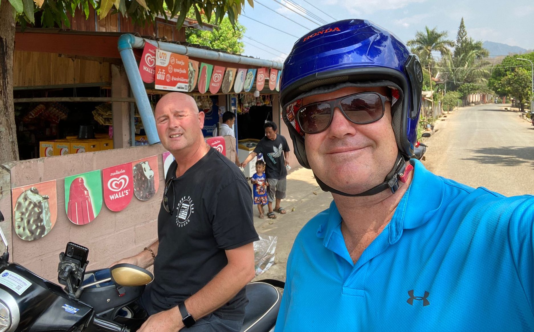 Two men taking a selfie on a motorcycle in a street setting, with a shop in the background and a child walking nearby.