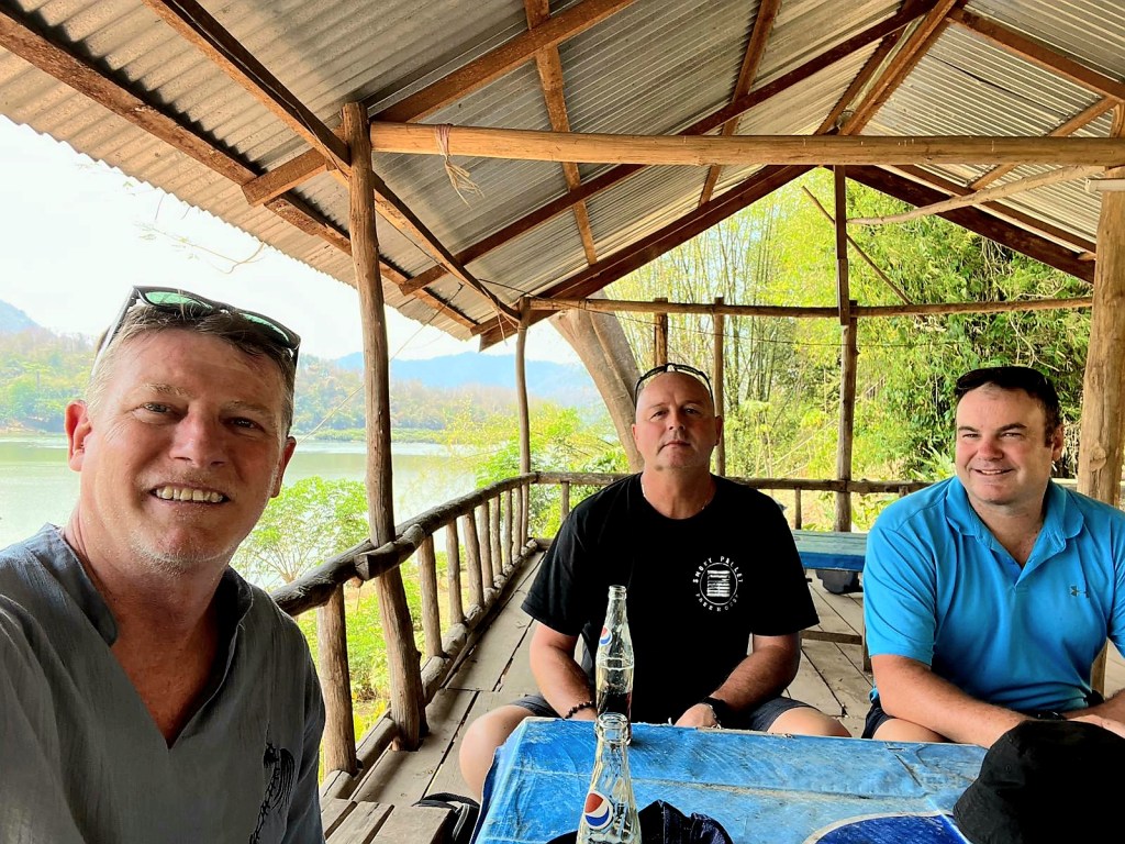 A group of three men sitting at a wooden table under a covered structure near a lake, with mountains and greenery in the background.