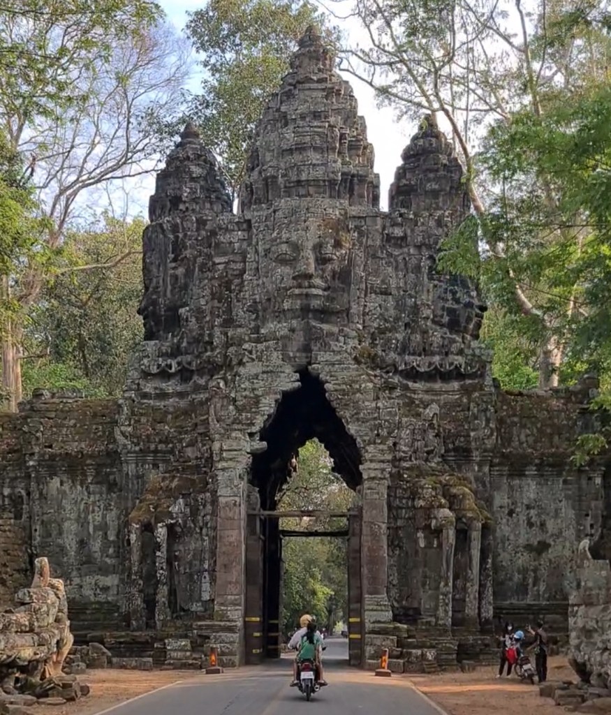 Historic stone gate with faces carved into it, surrounded by trees, with a motorcycle passing through.