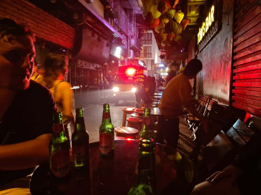 A busy street scene at night with people socializing outside a bar. Several green beer bottles are visible on tables, illuminated by red and purple lights. A vehicle with flashing lights is seen in the background, adding to the vibrant atmosphere.