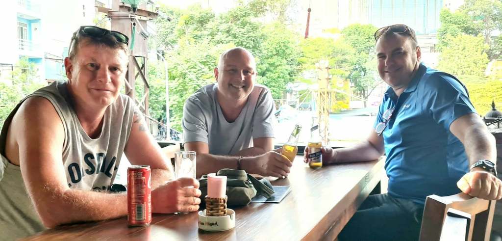 Three men sitting at a wooden table in a cafe, enjoying drinks and smiling at the camera.