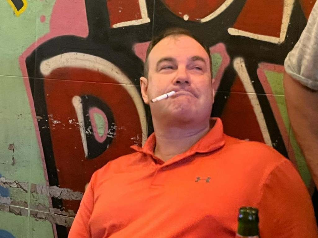 A man sitting against a colorful graffiti wall, wearing an orange polo shirt, with a cigarette in his mouth and a bottle of beer in front of him.