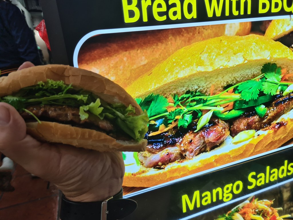 A person holding a sandwich filled with greens and meat, in front of a colorful menu sign advertising 'Bread with BBQ' and 'Mango Salads.'