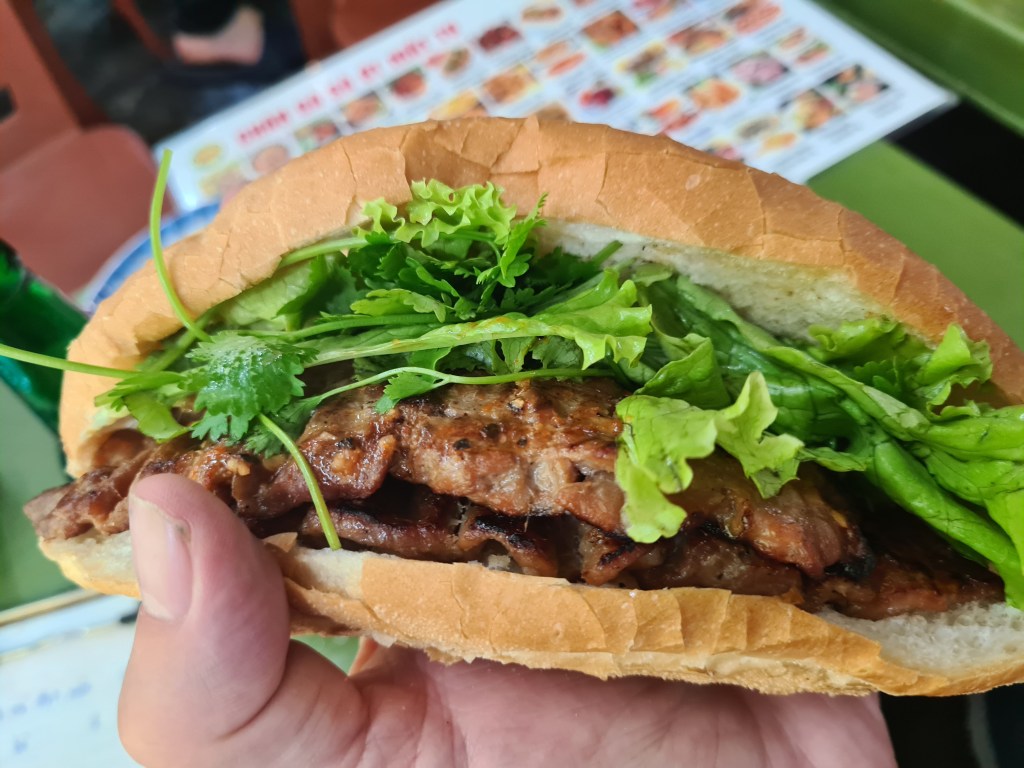 A hand holding a Vietnamese banh mi sandwich filled with grilled meat, fresh lettuce, and cilantro, with a green bottle and a menu in the background.