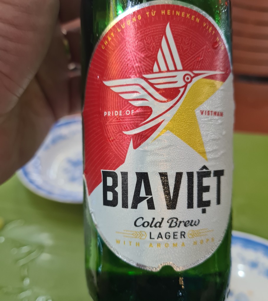 Close-up of a Bia Viet Cold Brew Lager bottle, featuring a red and yellow label with a bird logo, held by a hand against a green table.