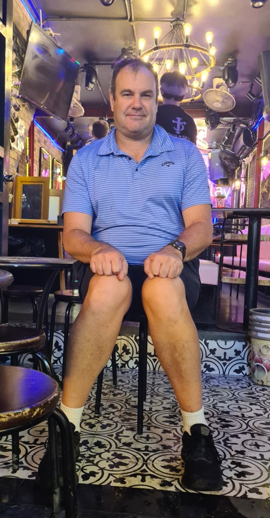 A man sitting on a chair in a dimly lit bar, wearing a blue polo shirt and black shorts. The background features decorative lighting and a patterned floor.