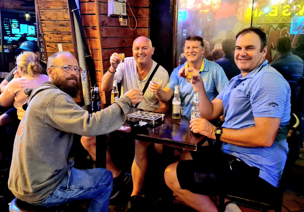 Group of four men sitting at a table in a bar, holding up drinks and smiling. They are surrounded by a casual atmosphere with wooden decor.