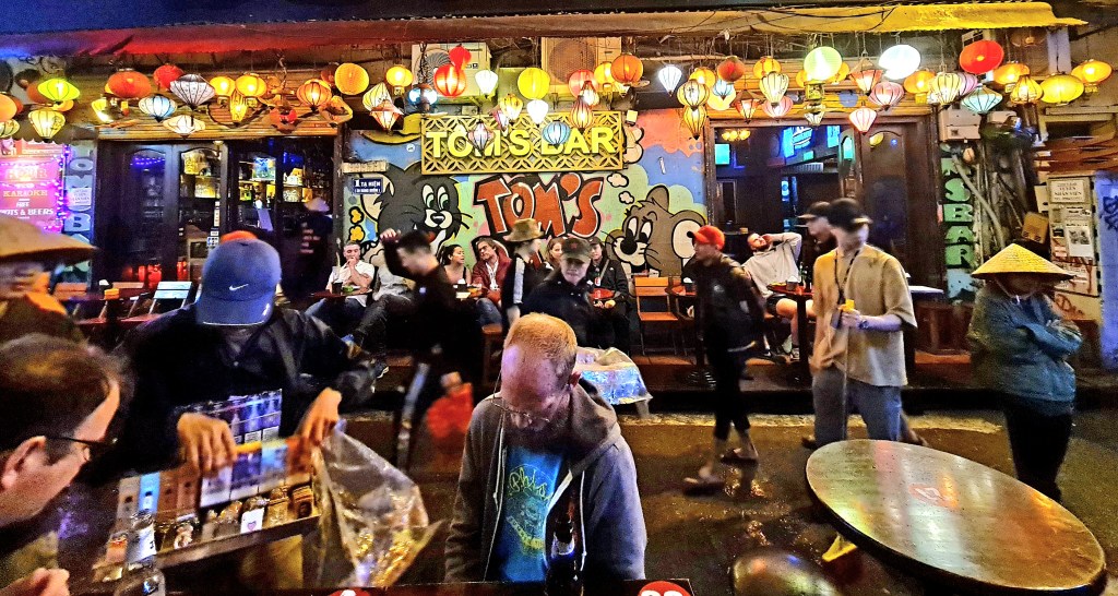 A crowd of people interacting in front of a vibrant bar decorated with colorful lanterns and graffiti. Some individuals are seated while others are busy, creating a lively atmosphere.