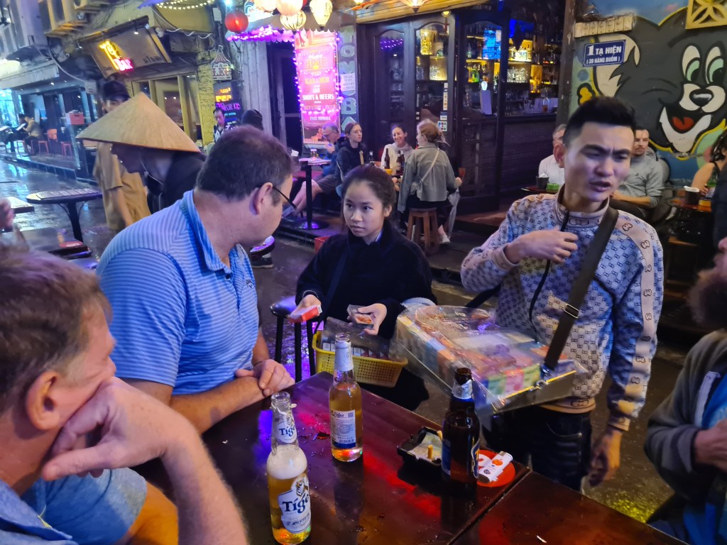 A bustling street scene at night with people sitting at tables outside bars. A man in a blue shirt is talking to a young woman holding a tray of items, while another man in a patterned shirt stands nearby. Several drinks are on the table, and there are colorful lights and decorations in the background.