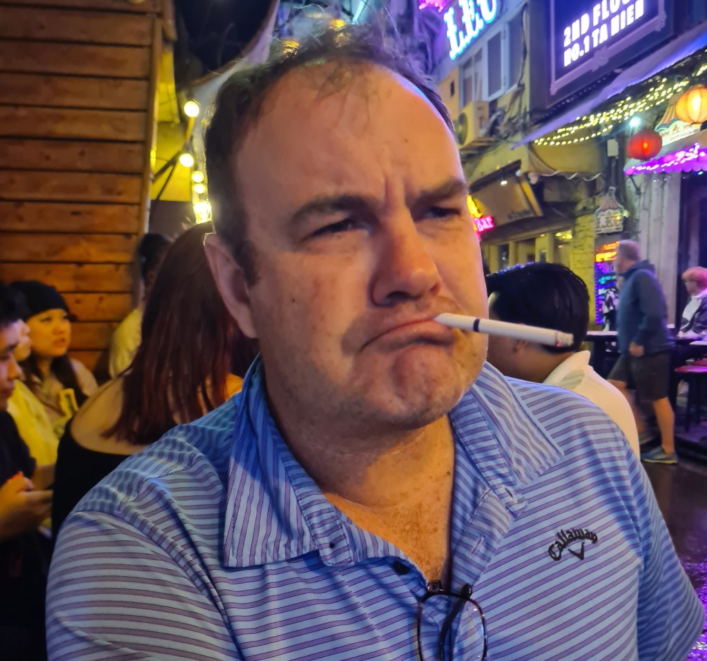 A man with a cigarette resting in his mouth, looking pensive, in a vibrant nightlife setting with colorful lights in the background.