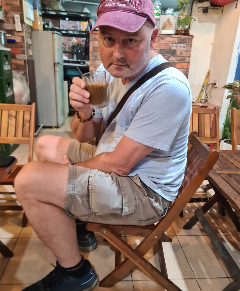 A man sitting on a wooden chair in a casual setting, holding a glass of brown beverage and looking pensive.