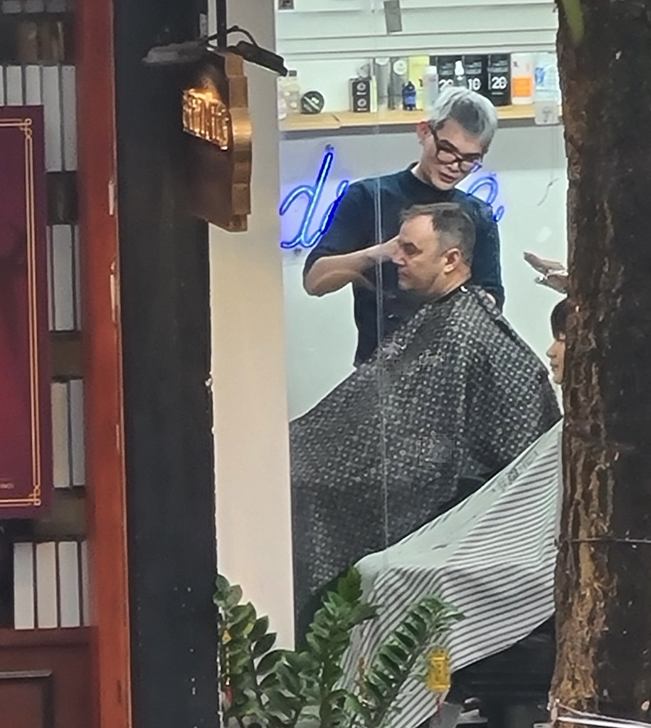 A barber with gray hair and glasses cuts the hair of a seated man draped in a black and white patterned cape inside a barbershop.