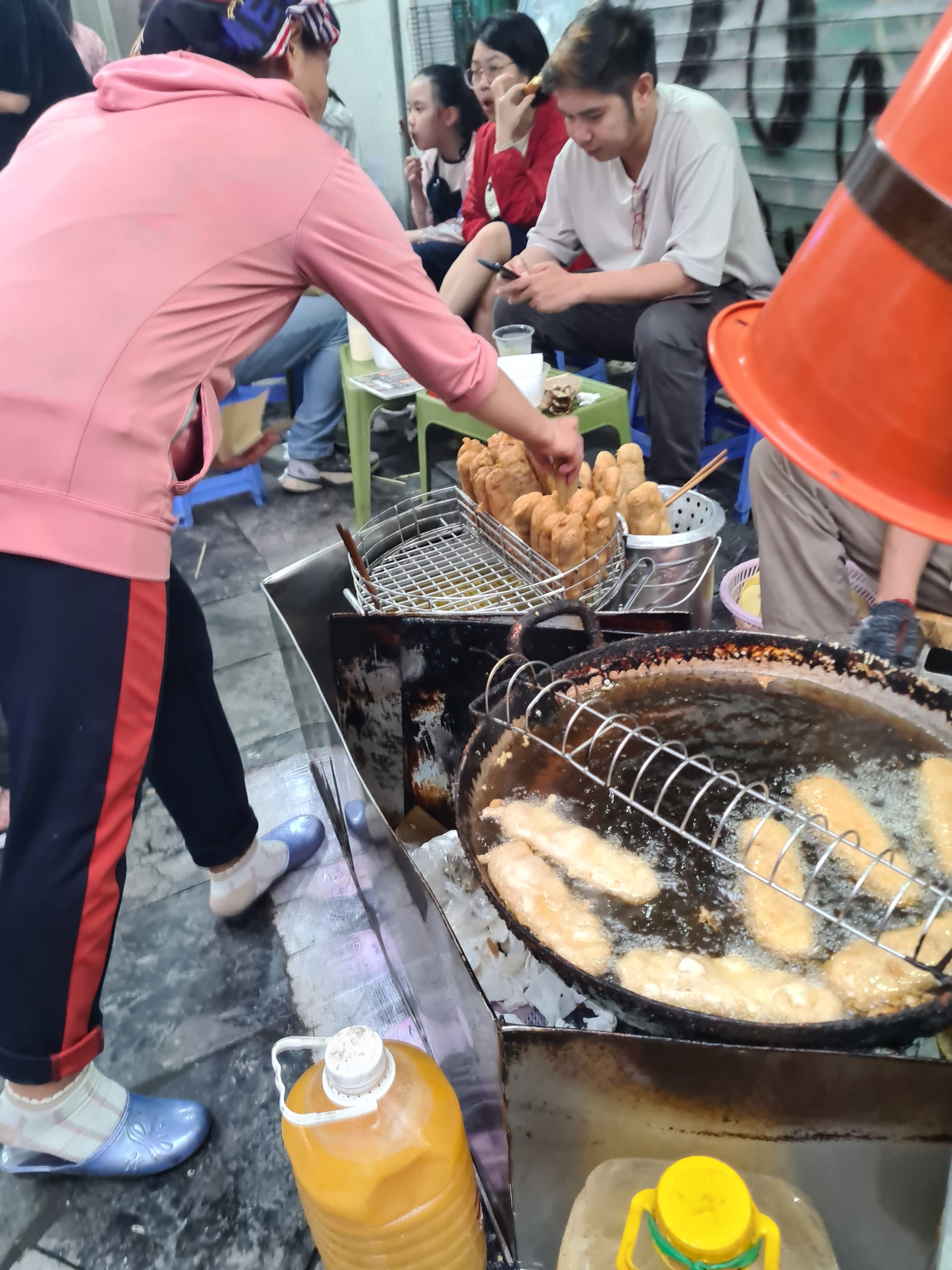 A street vendor frying food in oil, with other customers seated nearby enjoying snacks. The vendor wears a pink sweatshirt and is handling food on a skewer. Bottles of oil are visible beside the fryer.