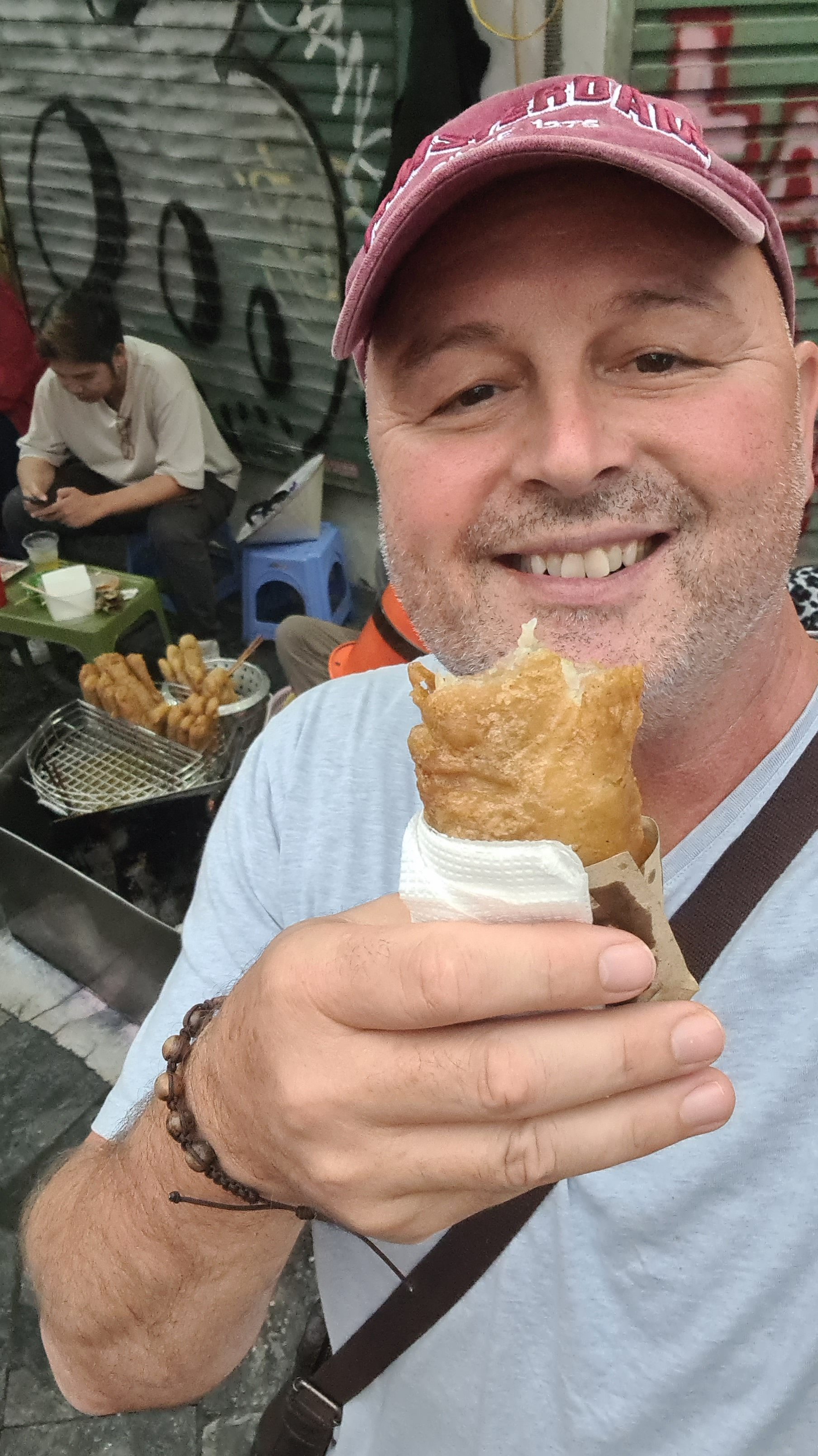 A smiling man holding a fried snack in a paper wrap while taking a selfie in a street food setting, with others sitting in the background.