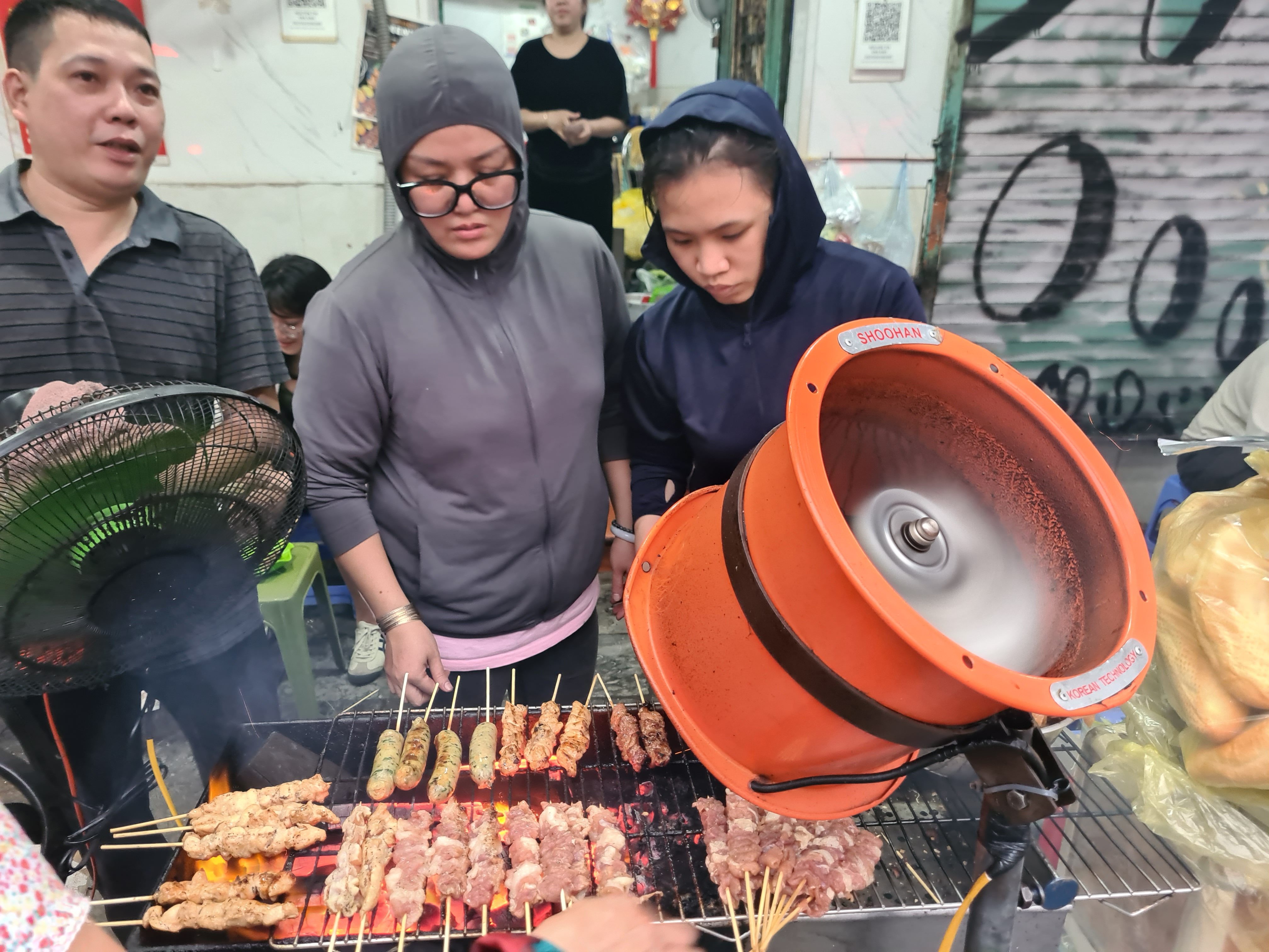 Several people grilling skewered meats over an open flame with a fan to disperse smoke, in a bustling street food setting.
