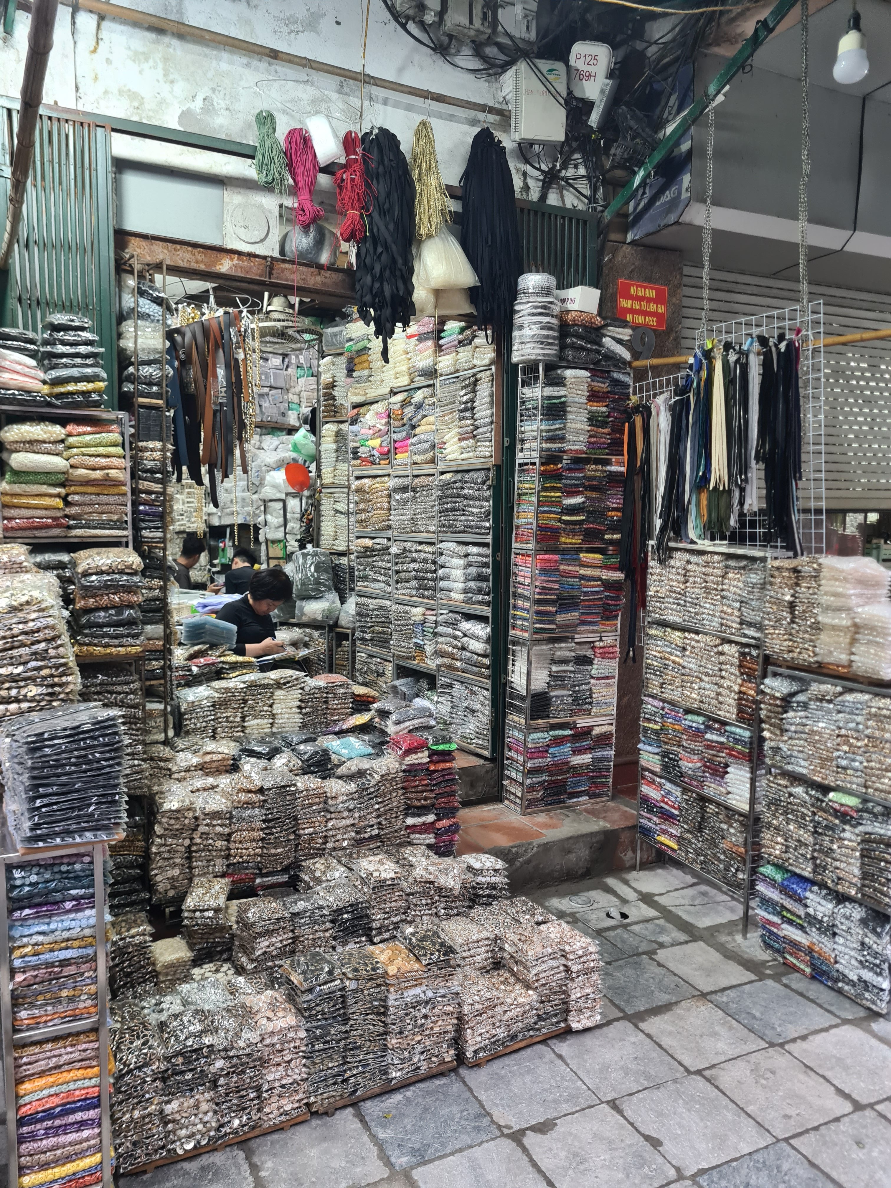 A bustling fabric market filled with stacks of colorful rolls of fabric and spools of thread, organized on shelves and tables, with a vendor sitting in the background.