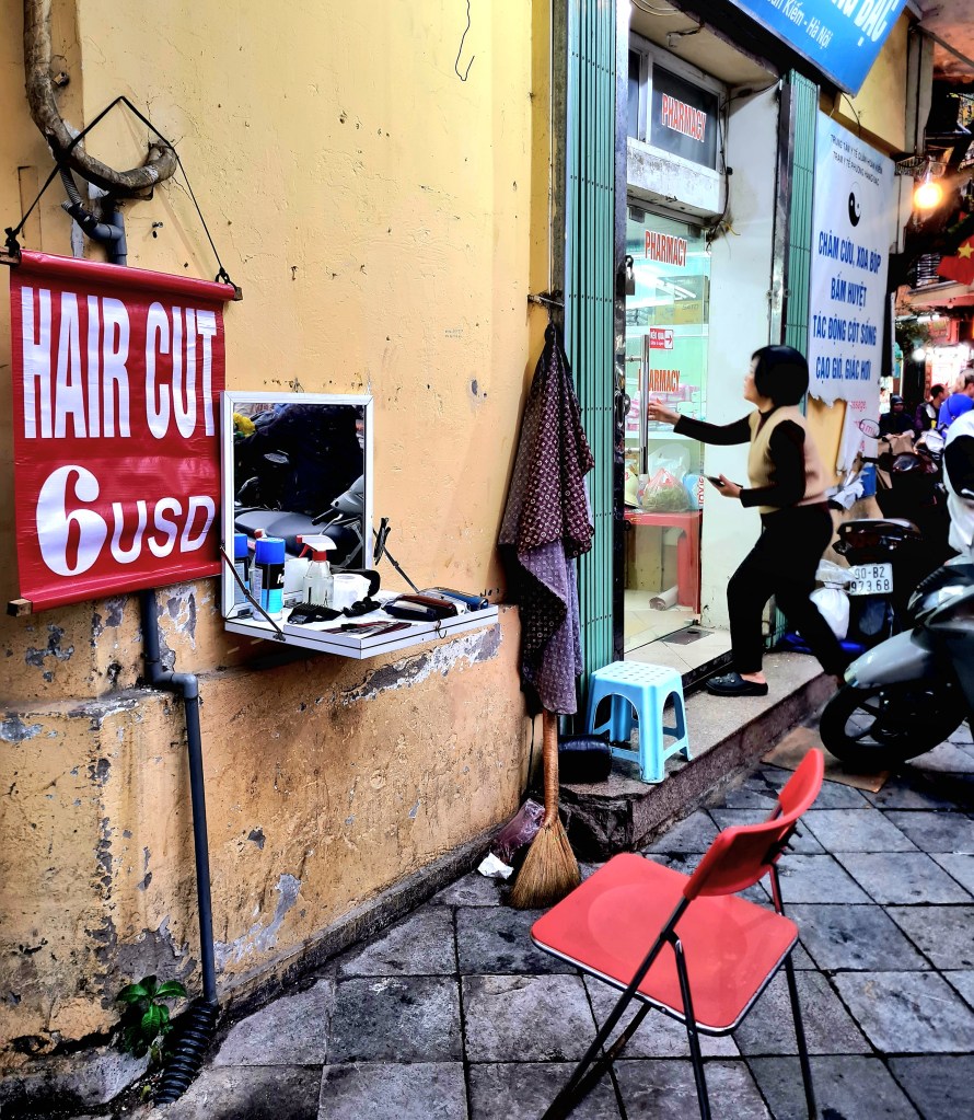Outdoor haircutting station with a sign reading 'HAIR CUT 6 USD', featuring a mirror, hair tools, a red chair, and a broom on the ground, adjacent to a pharmacy entrance.