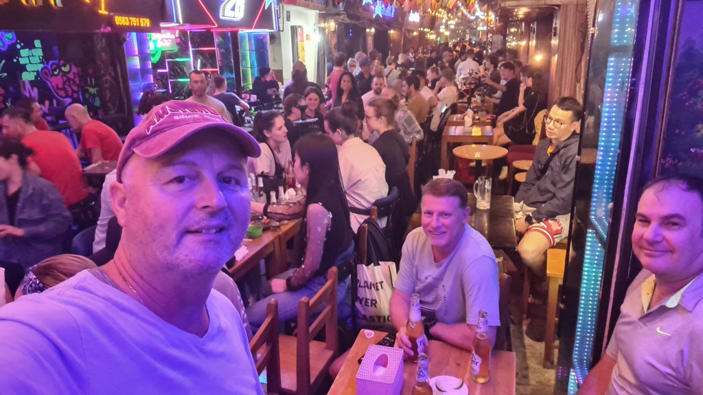 A crowded bar scene filled with people enjoying drinks and socializing, with three men in the foreground smiling at the camera.