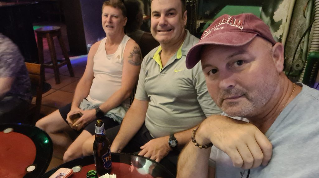 Three men sitting at a table in a bar, smiling and posing for a selfie. One man is wearing a white tank top, another is in a gray shirt, and the third is wearing a maroon cap. There are drinks on the table.
