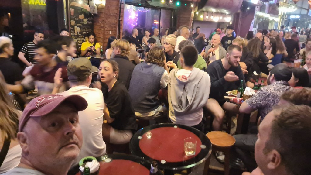 A crowded bar scene with diverse groups of people socializing, eating, and drinking. The foreground features a man taking a selfie, while others are engaged in conversations, with food and drinks present on tables.