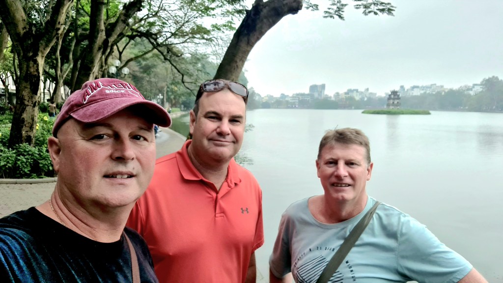 Three people standing by a serene lake surrounded by trees, with a distant island and city skyline in the background.