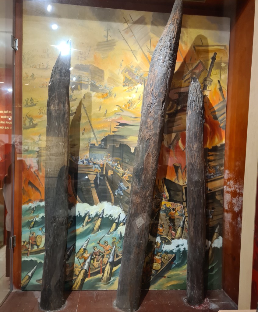 Three large wooden logs displayed in a glass case, with a vibrant mural in the background depicting a historical battle scene involving ships and warriors.