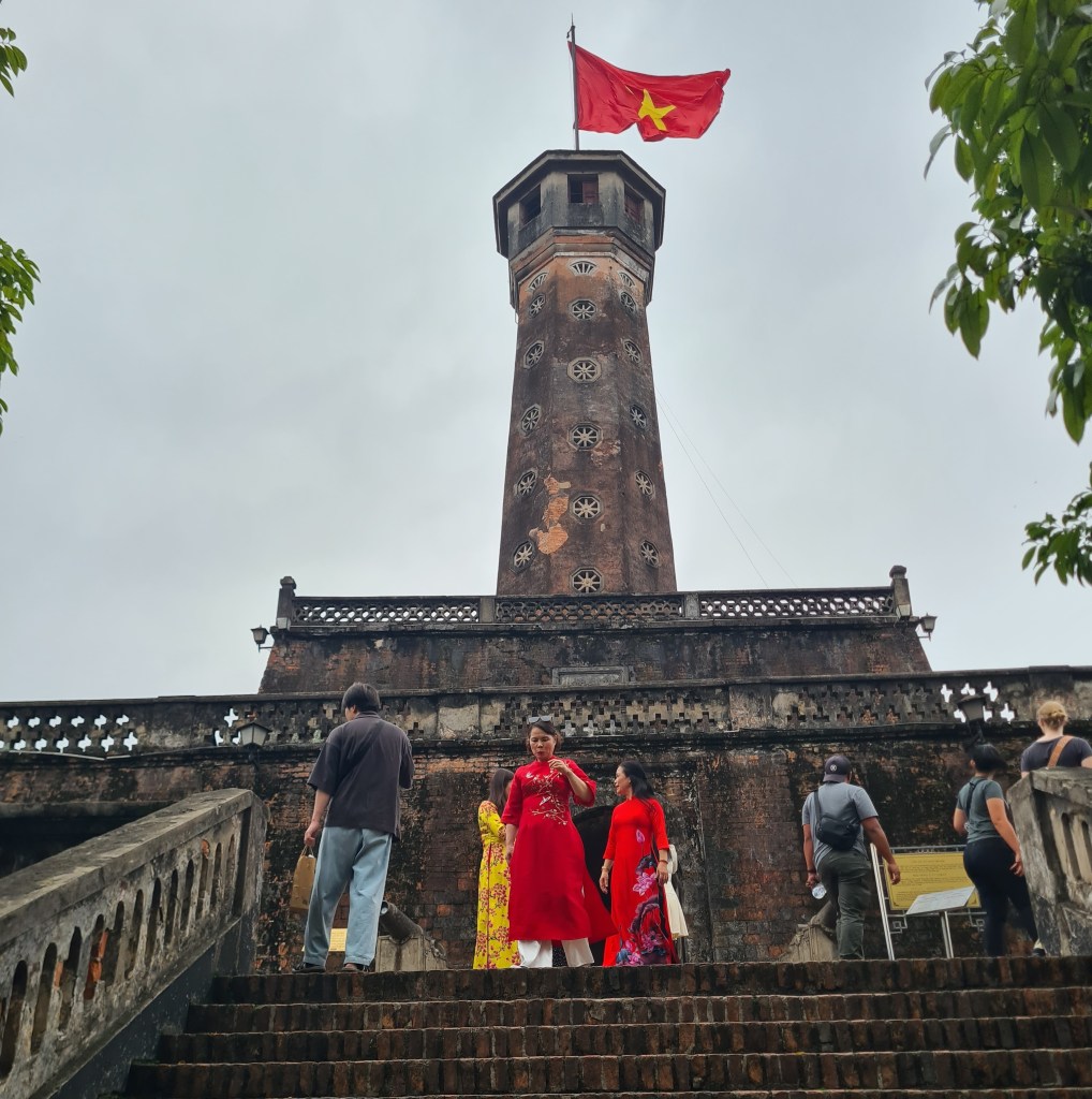 People walking down the steps towards the Imperial Citadel tower in Hanoi, Vietnam, with a Vietnamese flag waving atop.