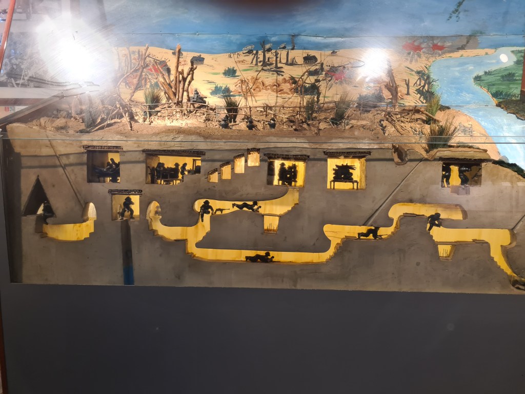 A detailed diorama depicting a military scene with miniature figures positioned in various locations, showcasing battle preparations and landscape features such as hills, water, and vegetation.