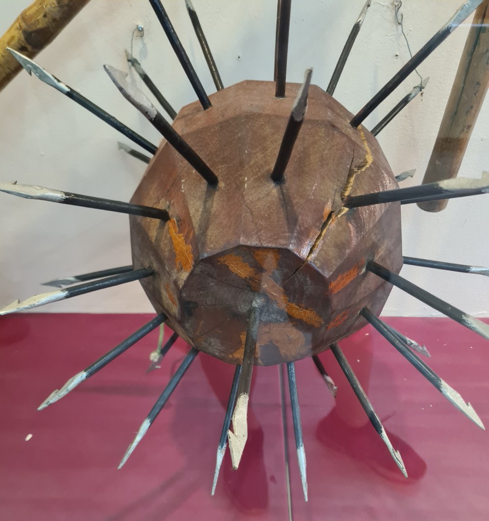 An antique spiked weapon resembling a spherical shield with multiple sharp metal spikes protruding from its surface, displayed on a red tabletop.